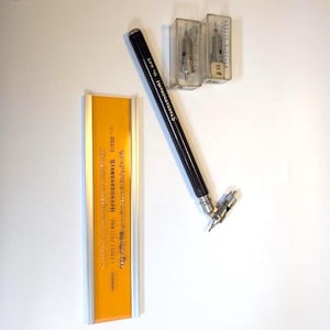 May include: A black Standardgraph No. 445 technical pen with a metal tip, an orange and silver ruler labeled Standardgraph 203/2, and two clear plastic cases containing metal pen nibs, one of which is labeled with the number 55.9.