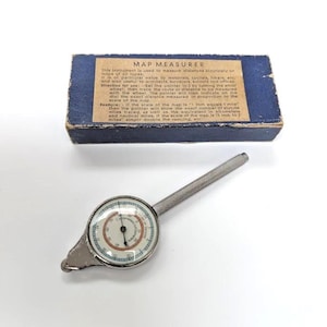 May include: A silver-toned metal map measurer with a circular dial featuring black, red, and blue markings, attached to a textured cylindrical handle. It is accompanied by a rectangular blue storage box with a tan label titled MAP MEASURER.
