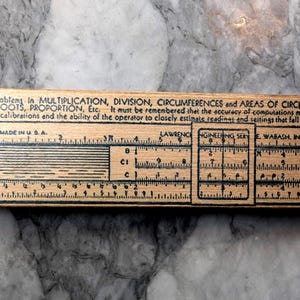May include: A vintage wooden slide rule with black printed markings and text, including "MADE IN U.S.A." and instructions for calculations. The slide rule is designed for solving problems in multiplication, division, and other mathematical functions.
