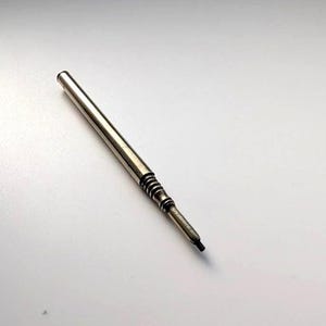 Op de afbeelding: Een zilverkleurige metalen penvulling met een gladde cilindrische behuizing die taps toeloopt in een geribbeld gedeelte bij de punt. Het uiteinde is smal en bevat een kleine zwarte punt, terwijl de schacht recht en gepolijst is.