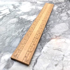 Könnte beinhalten: Ein hölzernes Lineal mit geätzten Markierungen und Zahlen. Das Lineal ist hellbraun und etwa 30 cm lang. Die Markierungen sind schwarz und gleichmäßig über die Länge des Lineals verteilt.