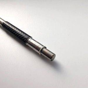Puede incluir: Un instrumento de escritura con un cuerpo negro de patrón de espiga texturizado y una punta y tapa final de metal plateado. El cuerpo cilíndrico pasa de la empuñadura texturizada a una sección plateada lisa y pulida con un pequeño anillo.