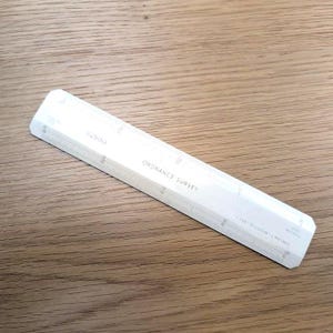May include: A white Ordnance Survey scale ruler with measurements and text. The ruler is angled on a wooden surface. The text "Ordnance Survey" is visible. The scale is marked with numbers and divisions.