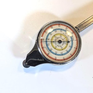 May include: A vintage map measurer with a silver body and a circular dial. The dial has markings for miles, kilometres, and nautical miles, with a needle indicating a distance. The dial features a white, yellow, blue, and red colour scheme.