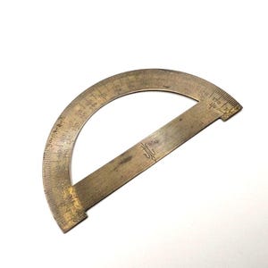 Puede incluir: Un transportador de latón antiguo con una escala semicircular y un borde recto. El transportador tiene marcas de grados grabadas y un emblema decorativo. El latón tiene una pátina cálida y envejecida, lo que sugiere que es una herramienta de dibujo antigua.