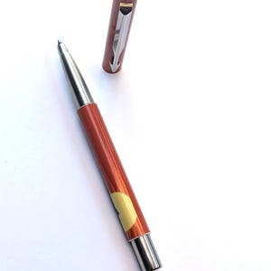 May include: A pen with a metallic silver tip and a reddish-brown barrel. The pen's cap is off and placed next to the pen. The pen has a silver clip and a small yellow accent. The pen is on a white background.