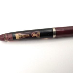 Könnte beinhalten: Burgunderfarbener Füllfederhalter mit schwarzem Band und silberner Zierleiste. Der Schaft des Stifts zeigt ein dekoratives Bild einer Flasche und einer Person. Der Stift hat eine transparente Spitze und ist vor einem weißen Hintergrund platziert.