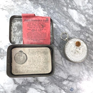 Può includere: Un kit di fotografia d'epoca con una scatola di metallo contenente Rapid Deadmatch Paper e un esposimetro circolare. La scatola ha testo e un inserto di carta rossa. L'esposimetro ha un quadrante con numeri e testo.