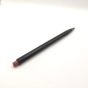 Puede incluir: Un portaminas negro con una goma de borrar roja. El lápiz tiene un cuerpo hexagonal y una punta metálica. La goma es cilíndrica y de color rojo oscuro. El lápiz está sobre una superficie blanca.