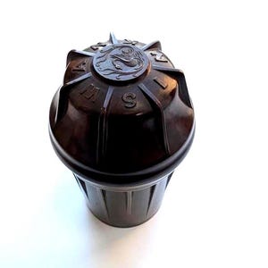Peut inclure: Un récipient cylindrique marron foncé avec un extérieur nervuré et un couvercle circulaire. Le couvercle présente un emblème central en relief représentant un cygne, entouré des lettres S, W, A, N et N disposées en rayons entre les nervures.
