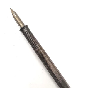 Op de afbeelding: Een vintage dip pen met een metalen penpunt en een donkerhouten handvat. De penpunt is zilverkleurig en puntig, terwijl het handvat een getextureerde, geribbelde grip heeft. De pen is ontworpen voor kalligrafie of tekenen, tegen een witte achtergrond.
