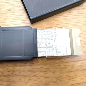 May include: A vintage slide rule with a black plastic frame and a white and beige measuring scale. The scale features various mathematical functions and measurements. The slide rule is partially extended, revealing its internal components.
