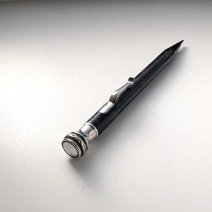 Puede incluir: Un portaminas negro con una tapa de metal plateado pulido y un clip de bolsillo a juego. El instrumento de escritura presenta una punta cónica y un cuerpo cilíndrico con un extremo superior redondeado y decorativo en tono plateado.