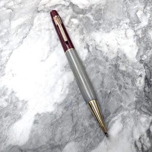 以下が含まれることがあります： ゴールドトーンのクリップが付いた深いバーガンディ色のキャップ、滑らかなグレーのバレル、磨かれたゴールドトーンの円錐形のペン先を備えたボールペン。このペンは、色分けされたセクションと金属製のアクセントが特徴の、洗練された円筒形のデザインです。