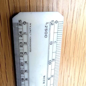 Può includere: Un righello di plastica bianca con segni e numeri neri, inclusi centimetri e frazioni. Il righello ha il testo "1/2500" e "WITH THE COMP" stampato sopra. Il righello è appoggiato su una superficie di legno.