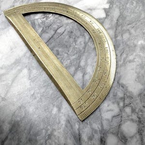 May include: A vintage brass protractor with degree markings from 0 to 170. The instrument has a straight edge and a curved, semi-circular scale. The brass has a warm, aged patina, suggesting its use over time.
