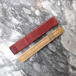 May include: A vintage wooden slide rule with a metal frame, next to a red leather case. The slide rule has various scales and markings. The case is rectangular and shows signs of wear. The items are on a marble surface.