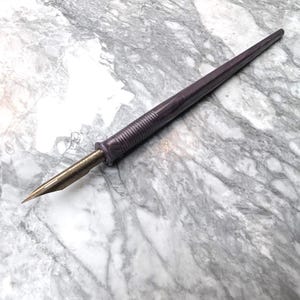 Puede incluir: Una pluma estilográfica morada con una plumilla metálica. El bolígrafo tiene un cuerpo largo y cónico y una sección de agarre texturizada. La plumilla es dorada y puntiaguda. El bolígrafo está diseñado para caligrafía y dibujo.
