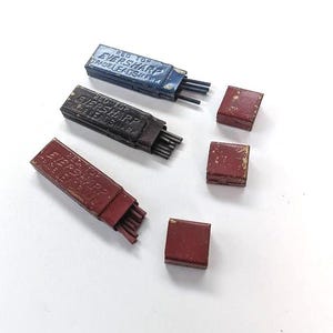 May include: Three rectangular metal containers for Eversharp Red Top leads, colored blue, black, and red, each containing thin graphite sticks. Three separate matching red metal lids are positioned to the right of the open containers.