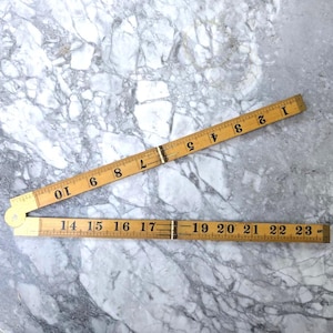 Puede incluir: Una regla plegable de madera vintage, con números negros sobre un fondo de madera clara. La regla está parcialmente abierta, mostrando medidas de 1 a 10 pulgadas y de 14 a 23 pulgadas (aproximadamente 36 a 58 cm).