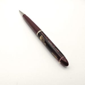 May include: A burgundy ballpoint pen with a silver tip and accents. The pen has a label with text that reads "Amontillado Medium Dry Sherry". The pen is on a white surface.
