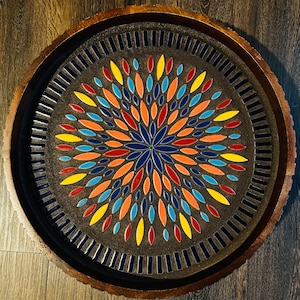 Könnte beinhalten: Ein runder dekorativer Tablett mit einem dunkelgrauen Zentrum, das ein farbenfrohes Blumenmuster aufweist. Das Design umfasst blaue, orangefarbene, gelbe und rote blattförmige Fliesen. Das Tablett hat einen dunkelbraunen Holzrand und einen helleren braunen Holzrand.
