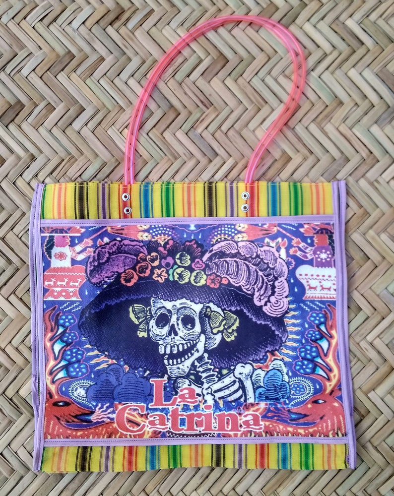 K&ouml;nnte beinhalten: Eine farbenfrohe Tragetasche mit einem gestreiften Design in Lila, Blau und Gelb. Die Tasche zeigt ein gro&szlig;es Bild eines Skeletts, das einen Hut mit Blumen tr&auml;gt und den Text "La Catrina".