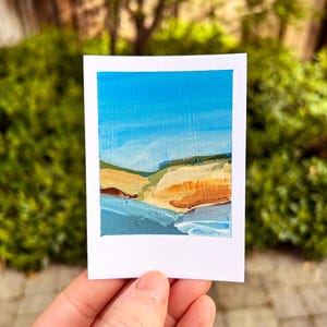 May include: A small, rectangular painting of a coastal landscape with a blue sky, sandy cliffs, and ocean waves. The painting is on a white card and held by a hand. The text "Wilder Ranch" is at the top.