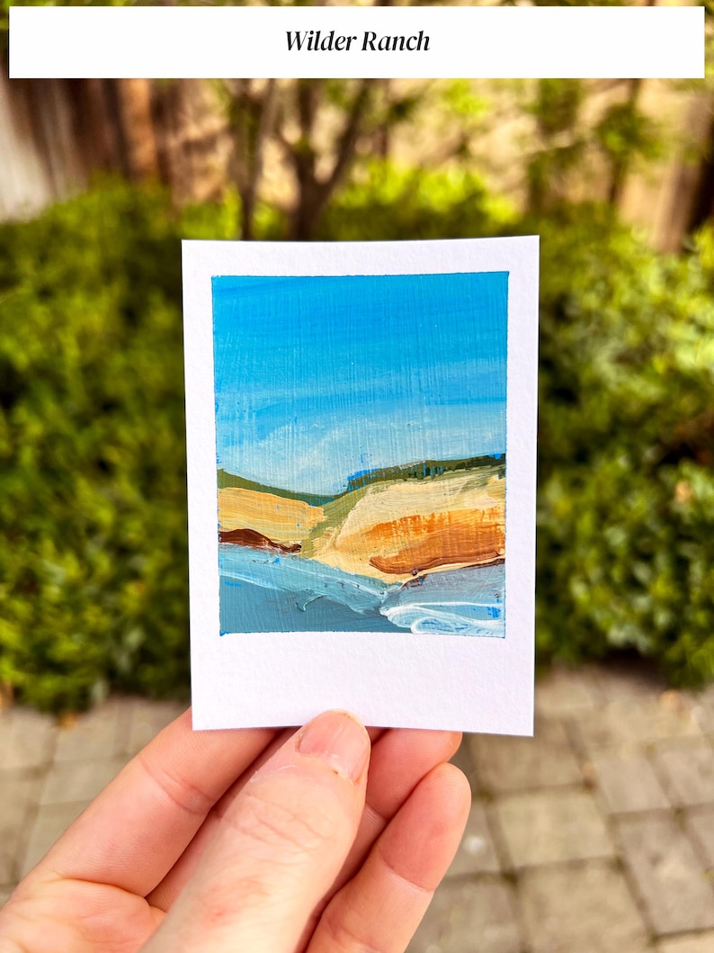 May include: A small, rectangular painting of a coastal landscape with a blue sky, sandy cliffs, and ocean waves. The painting is on a white card and held by a hand. The text "Wilder Ranch" is at the top.