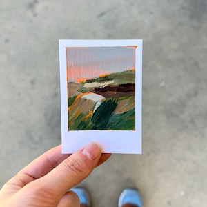 May include: A small landscape painting on a white card. The artwork features a sunset with orange and gray skies, green hills, and brown and white rock formations. The painting is held by a person.