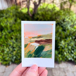 May include: A small, rectangular painting with the text "Point Reyes" at the top. The artwork features an abstract landscape with strokes of orange, green, brown, and beige. The painting is held in a person's hand against a backdrop of green foliage.