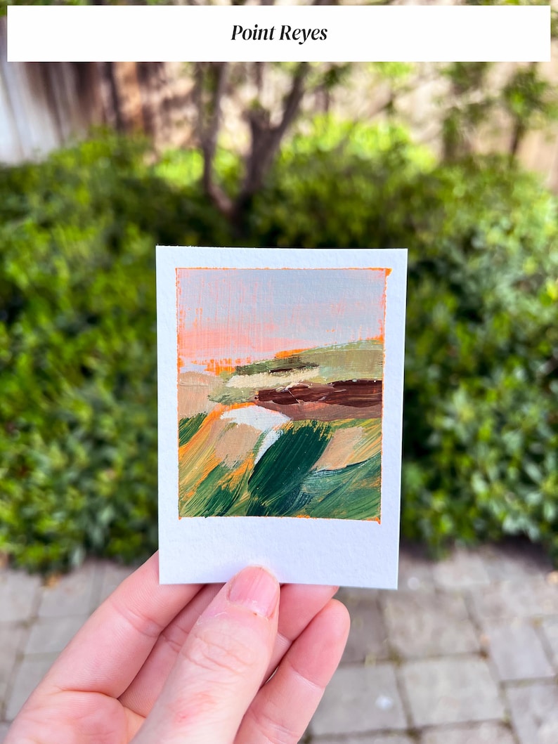 May include: A small, rectangular painting with the text "Point Reyes" at the top. The artwork features an abstract landscape with strokes of orange, green, brown, and beige. The painting is held in a person's hand against a backdrop of green foliage.