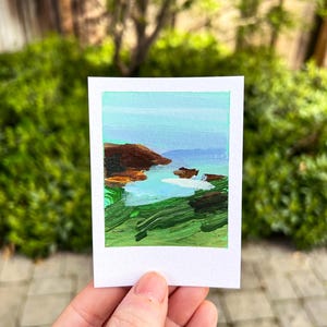 May include: A small, square painting titled "Sea Ranch" depicts a coastal landscape. The artwork features a blue sky, green grass, brown cliffs, and a body of water. The painting is held in a person's hand against a backdrop of green foliage.