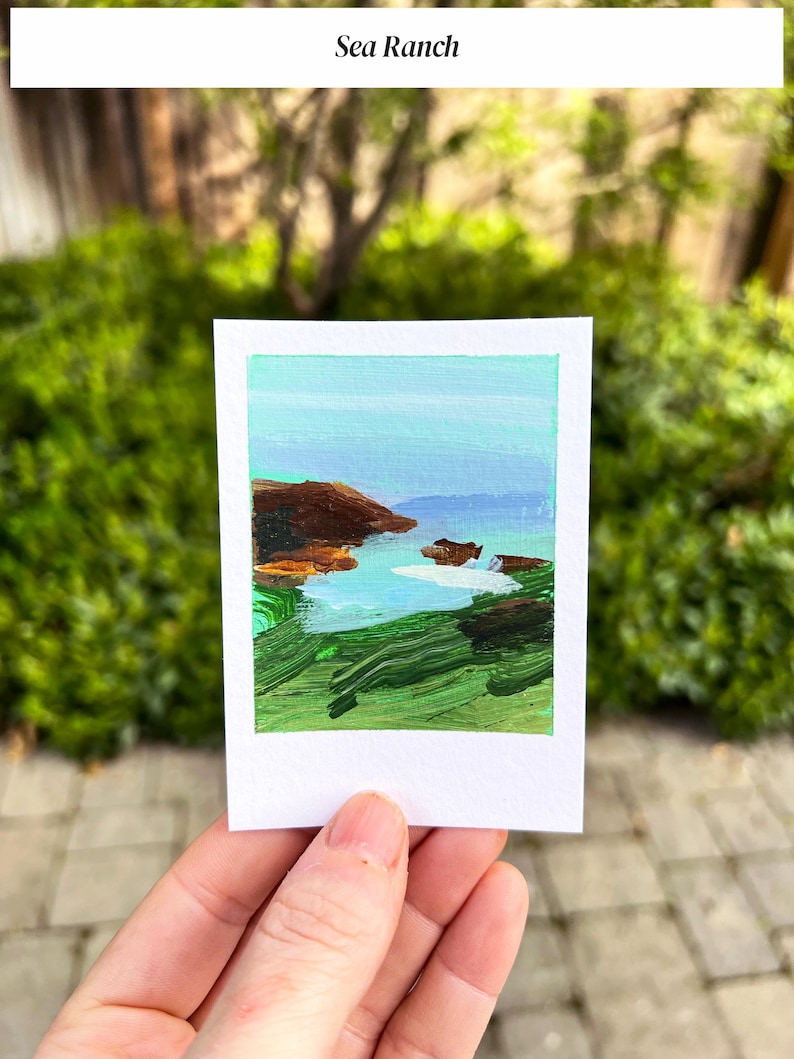 May include: A small, square painting titled "Sea Ranch" depicts a coastal landscape. The artwork features a blue sky, green grass, brown cliffs, and a body of water. The painting is held in a person's hand against a backdrop of green foliage.