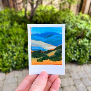 May include: A small, rectangular painting of a landscape with the text "Mt. Diablo" at the top. The artwork features blue mountains, green hills, and an orange sky. The painting is held in a person's hand against a backdrop of green foliage.