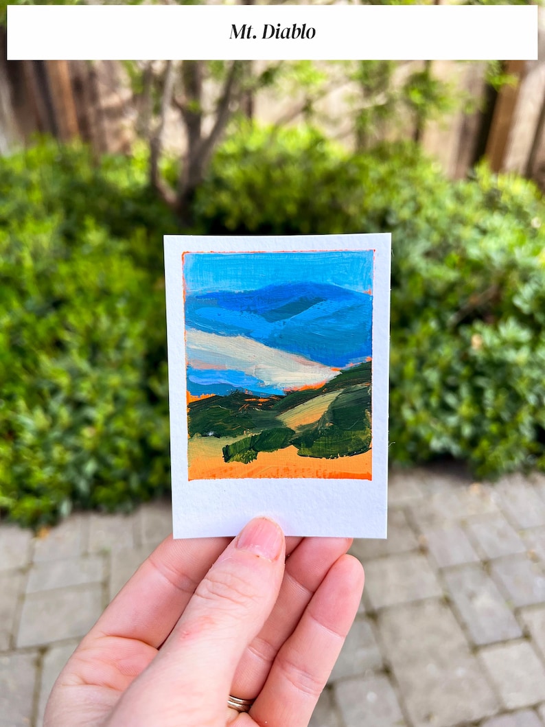 May include: A small, rectangular painting of a landscape with the text "Mt. Diablo" at the top. The artwork features blue mountains, green hills, and an orange sky. The painting is held in a person's hand against a backdrop of green foliage.