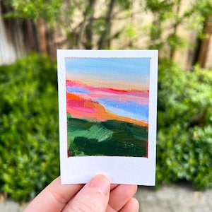 May include: A small, square painting on white paper with the text "Big Sur" at the top. The artwork depicts a landscape with a blue sky, pink and orange hues, and green terrain. The painting is held in a hand.