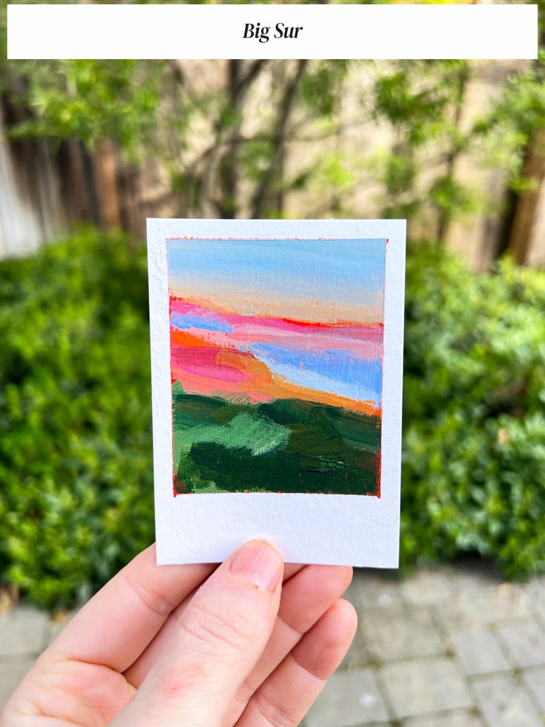 May include: A small, square painting on white paper with the text "Big Sur" at the top. The artwork depicts a landscape with a blue sky, pink and orange hues, and green terrain. The painting is held in a hand.