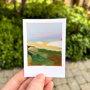 May include: A small, rectangular painting titled "Half Moon Bay" with a white border and an orange frame. The artwork depicts a landscape with green, yellow, and brown brushstrokes representing hills and fields, with a blue and pink sky.