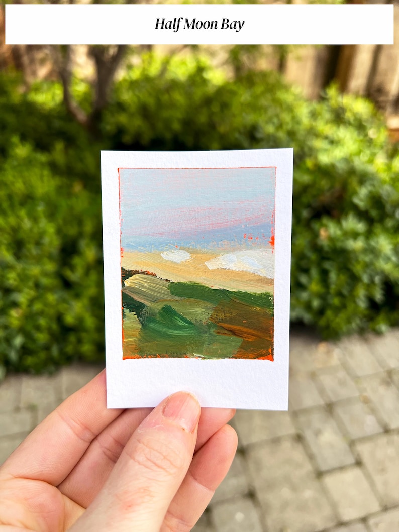 May include: A small, rectangular painting titled "Half Moon Bay" with a white border and an orange frame. The artwork depicts a landscape with green, yellow, and brown brushstrokes representing hills and fields, with a blue and pink sky.