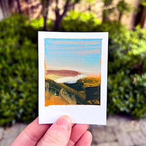 May include: A small, rectangular painting of a landscape with the text "Fort Funston" at the top. The artwork features a blue sky, a white and brown land mass, and dark green and orange brushstrokes. The painting is held in a person's hand.