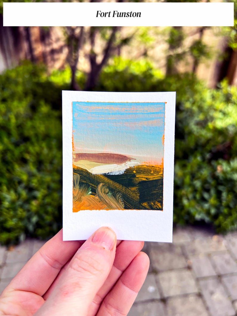 May include: A small, rectangular painting of a landscape with the text "Fort Funston" at the top. The artwork features a blue sky, a white and brown land mass, and dark green and orange brushstrokes. The painting is held in a person's hand.