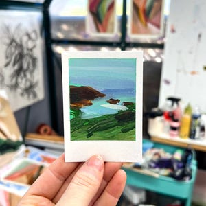 May include: A miniature landscape painting on a white paper, held in a hand. The artwork depicts a coastal scene with green, brown, and blue hues. The painting is in a Polaroid-style format, with a white border.