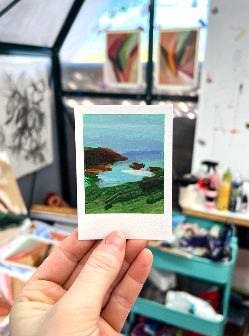 May include: A miniature landscape painting on a white paper, held in a hand. The artwork depicts a coastal scene with green, brown, and blue hues. The painting is in a Polaroid-style format, with a white border.