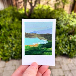 May include: A small landscape painting of Bodega Bay, featuring a turquoise bay, green hills, and a blue sky. The painting is on a white rectangular card, held by a hand, with the text "Bodega Bay" at the top.