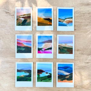 May include: Nine small landscape paintings on white frames. Each painting features a different abstract landscape with blue skies, green hills, and colorful brushstrokes. The paintings are arranged on a light brown wooden surface.