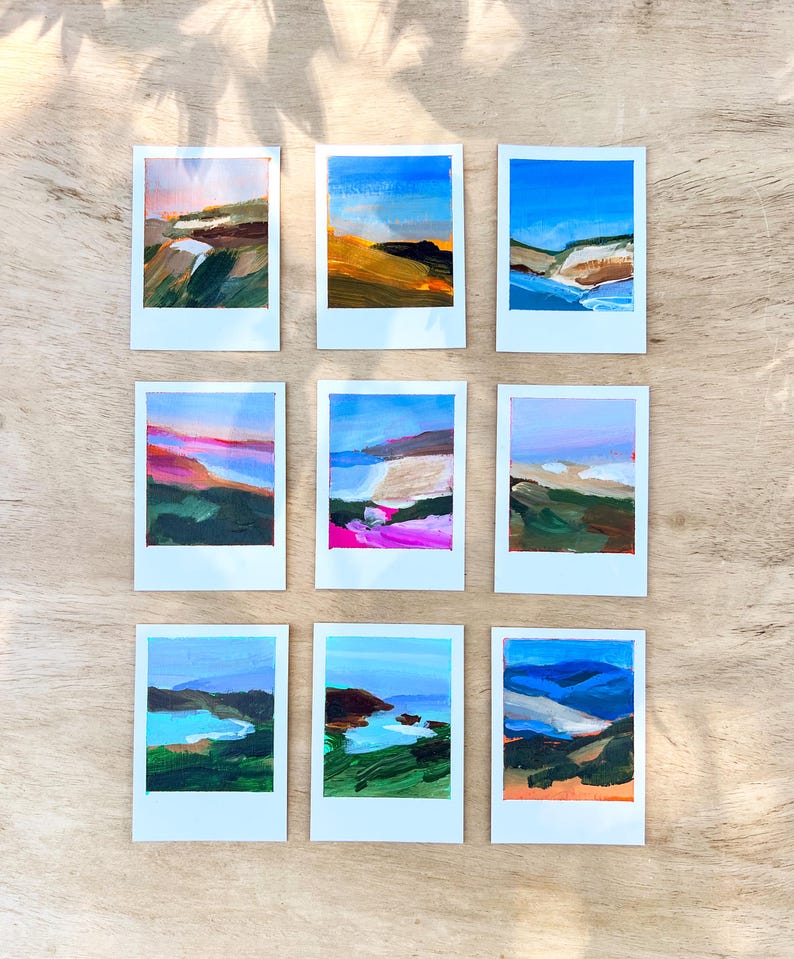May include: Nine small landscape paintings on white frames. Each painting features a different abstract landscape with blue skies, green hills, and colorful brushstrokes. The paintings are arranged on a light brown wooden surface.