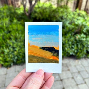 May include: A small, square painting titled "Marin Headlands" depicts a landscape with a blue sky and rolling hills in shades of yellow, orange, and black. The artwork is on a white card with a gold border, held against a blurred green background.