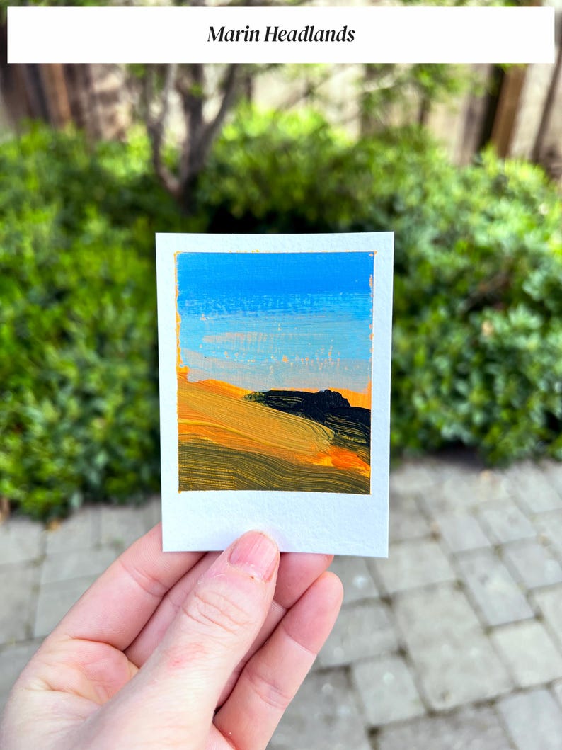 May include: A small, square painting titled "Marin Headlands" depicts a landscape with a blue sky and rolling hills in shades of yellow, orange, and black. The artwork is on a white card with a gold border, held against a blurred green background.