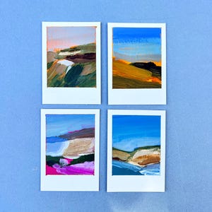 May include: Four small landscape paintings on white frames. Each painting features a different abstract landscape scene with brushstrokes of blue, green, brown, pink, and yellow. The paintings are arranged on a light blue surface.